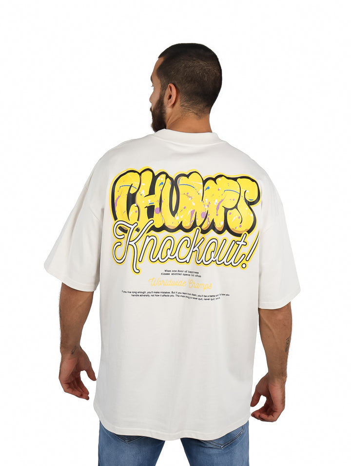 Hombre vistiendo playera oversize color arena con logo dorado en el pecho y pantalones mezclilla azules.