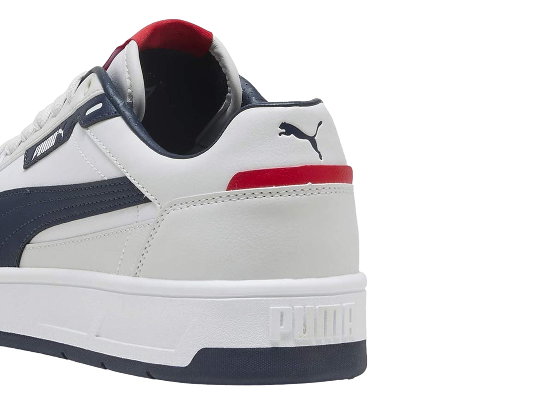 Tenis PUMA blancos con franja y suela azul marino y detalles rojos, vista lateral.