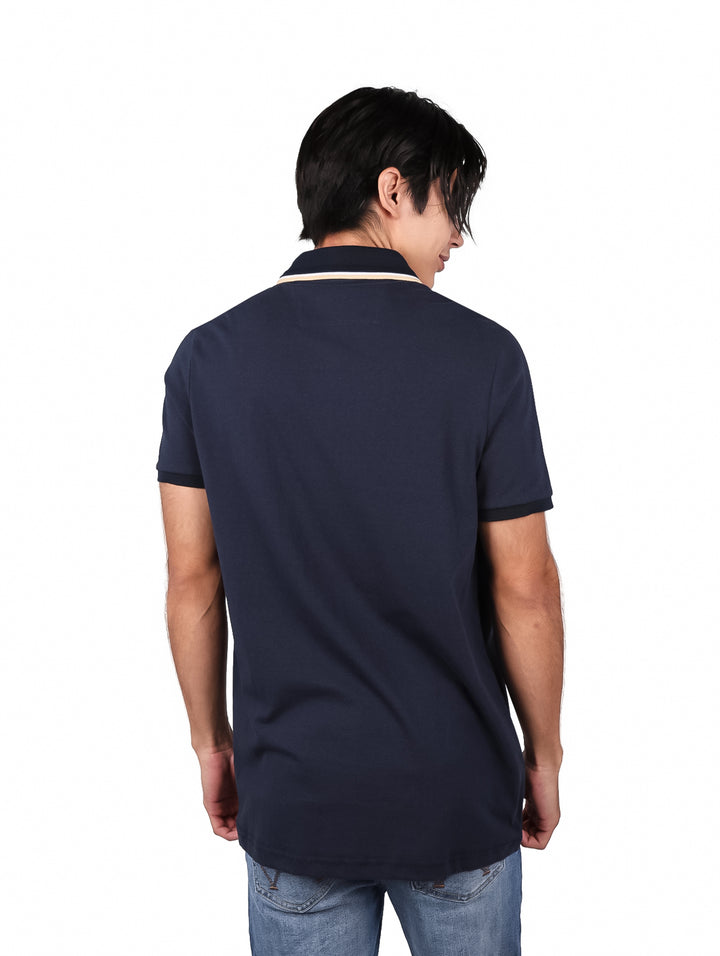 Hombre vistiendo una playera polo de color marino con detalle en el cuello y pantalones mezclilla azules.