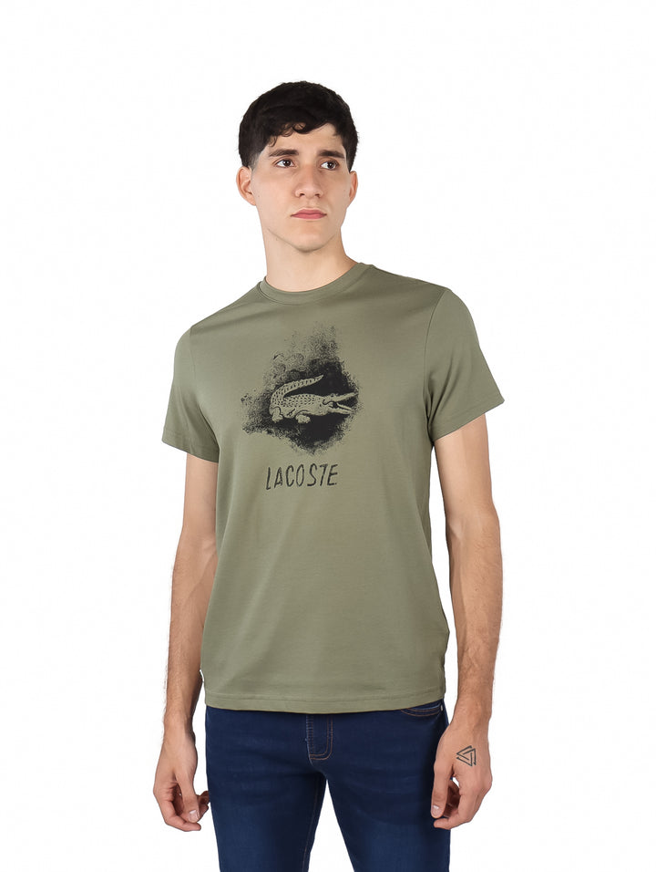 Hombre vistiendo camiseta de manga corta color kaki con gráfico de cocodrilo y texto Lacoste en el pecho.