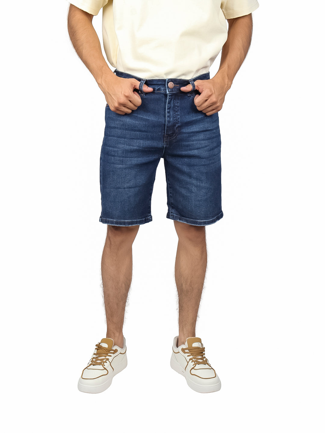 Hombre vistiendo bermudas de mezclilla color azul medio, camiseta crema y tenis blancos.