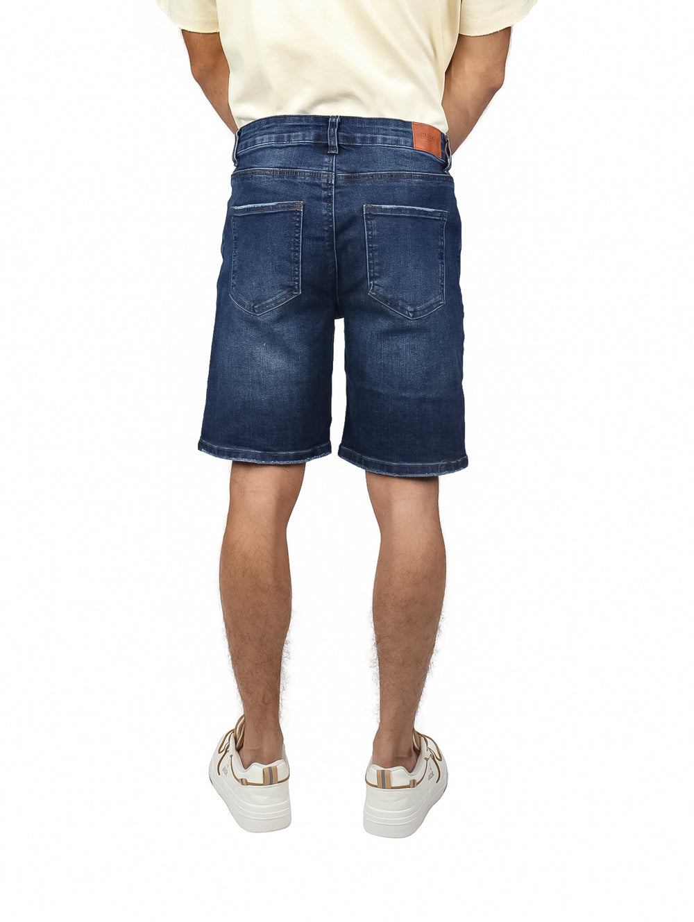 Hombre vistiendo bermudas de mezclilla color azul medio, camiseta crema y tenis blancos.