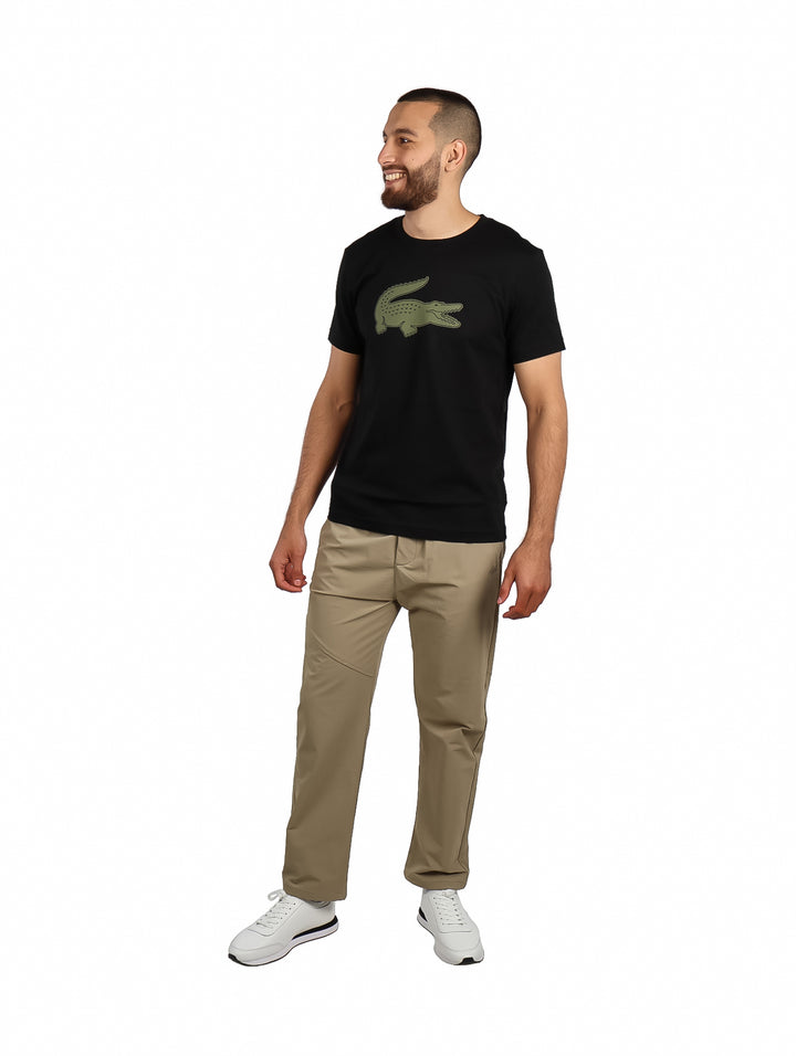 Hombre vistiendo camiseta negra de manga corta con un logotipo grande de cocodrilo en color kaki en el pecho.