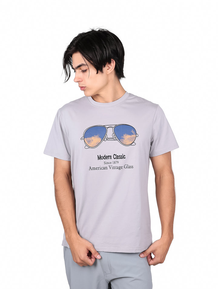 Hombre vistiendo playera gris de manga corta con estampado de lentes de sol y texto en el pecho.