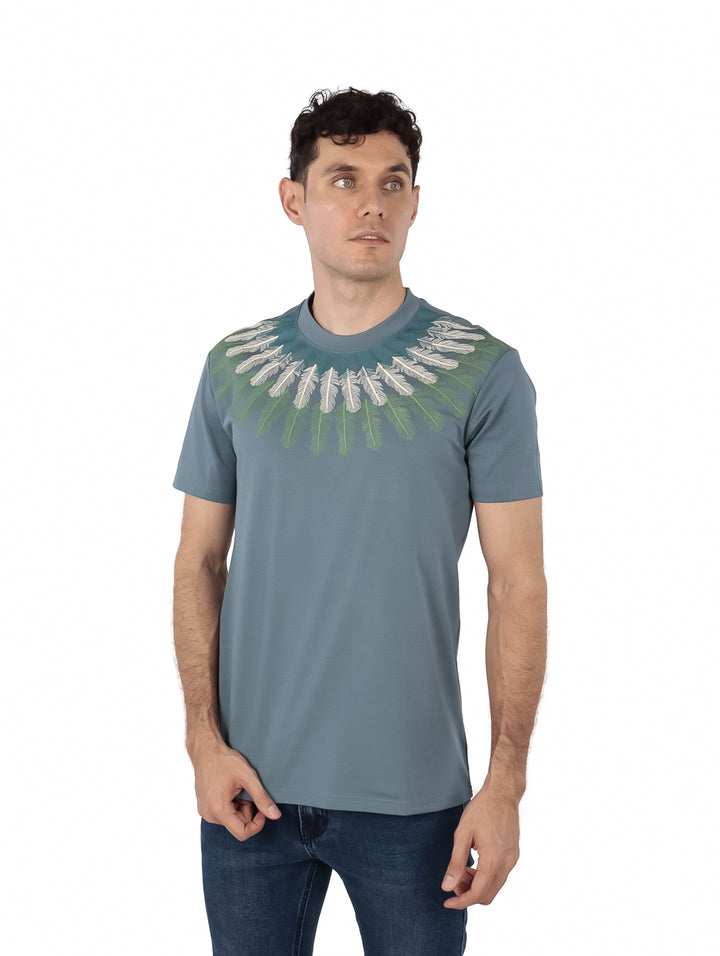 Hombre vistiendo playera color teal con estampado decorativo blanco y verde alrededor del cuello.