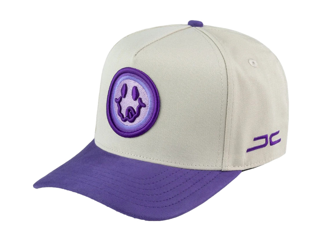 Gorra snapback color beige con visera y detalles en morado, incluye un parche circular frontal con diseño gráfico.