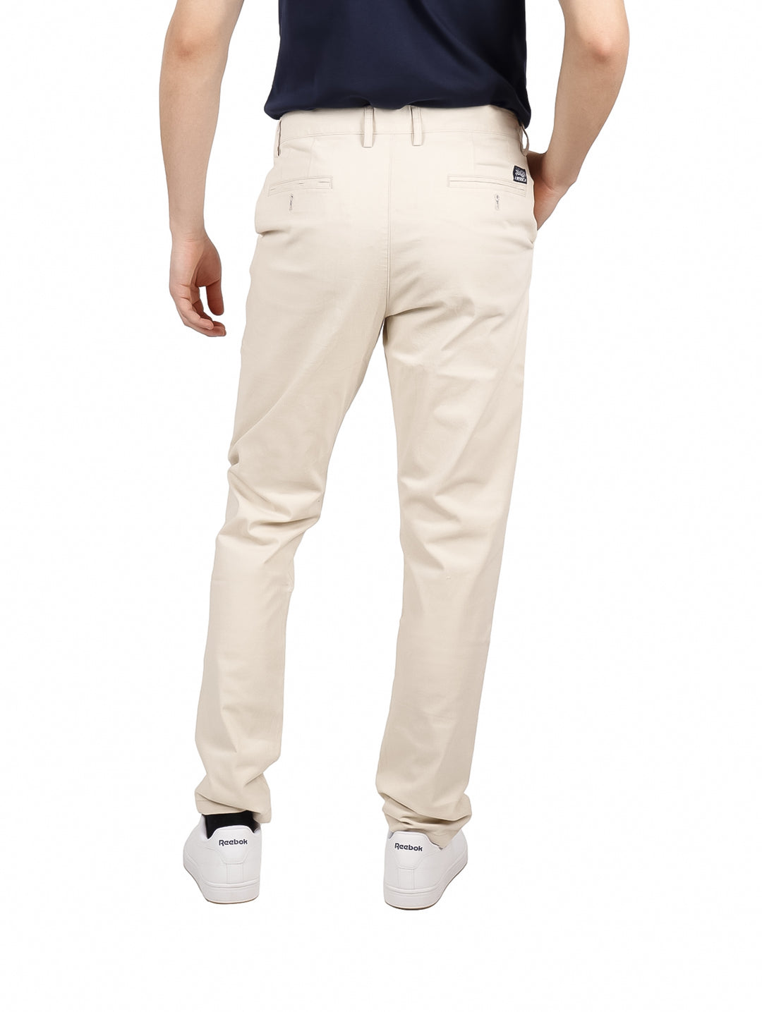 Hombre vistiendo pantalones chinos de color beige y tenis blancos sobre fondo blanco.