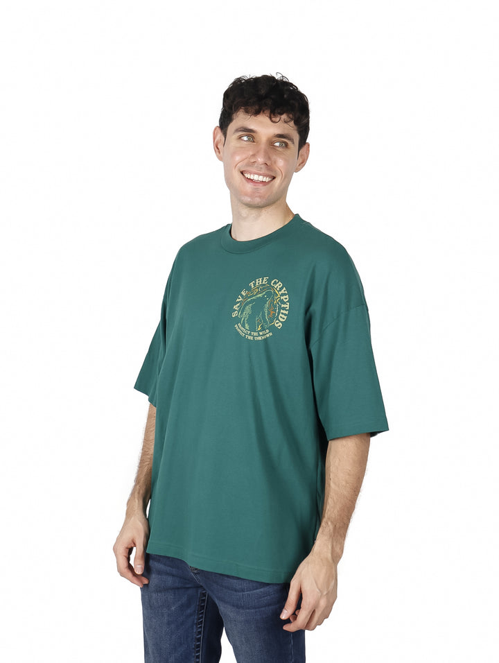 Hombre vistiendo playera oversized color verde botella con estampado amarillo en el pecho.