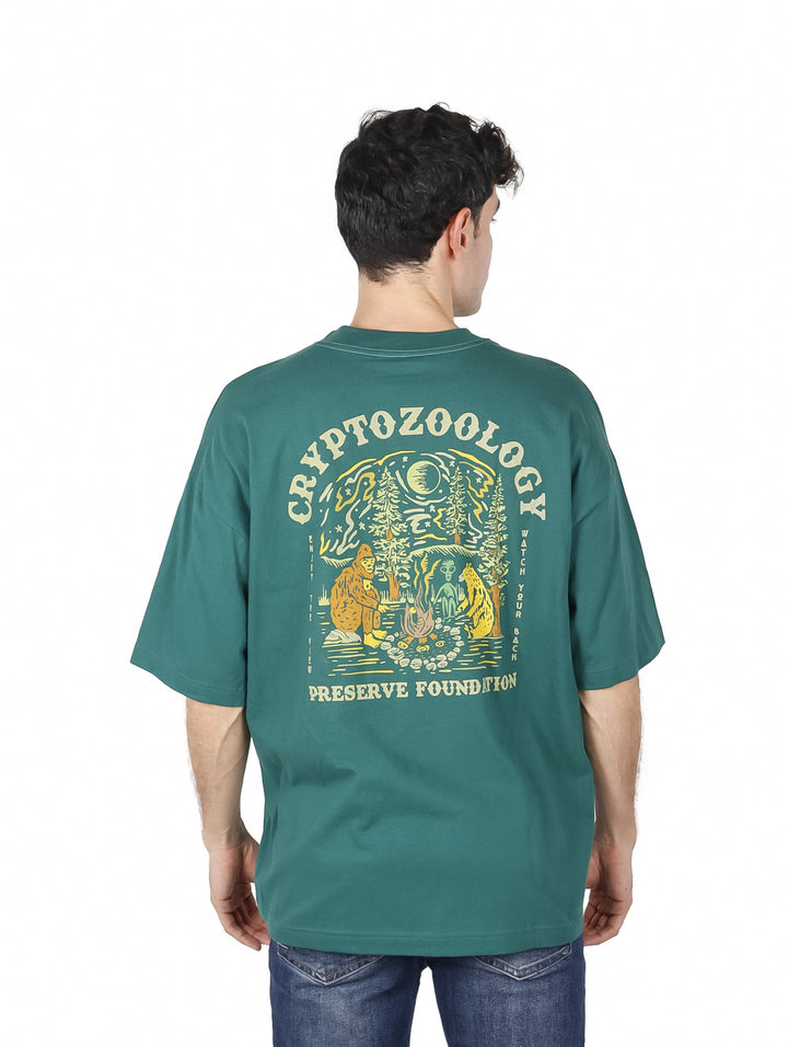 Hombre vistiendo playera oversized color verde botella con estampado amarillo en el pecho.