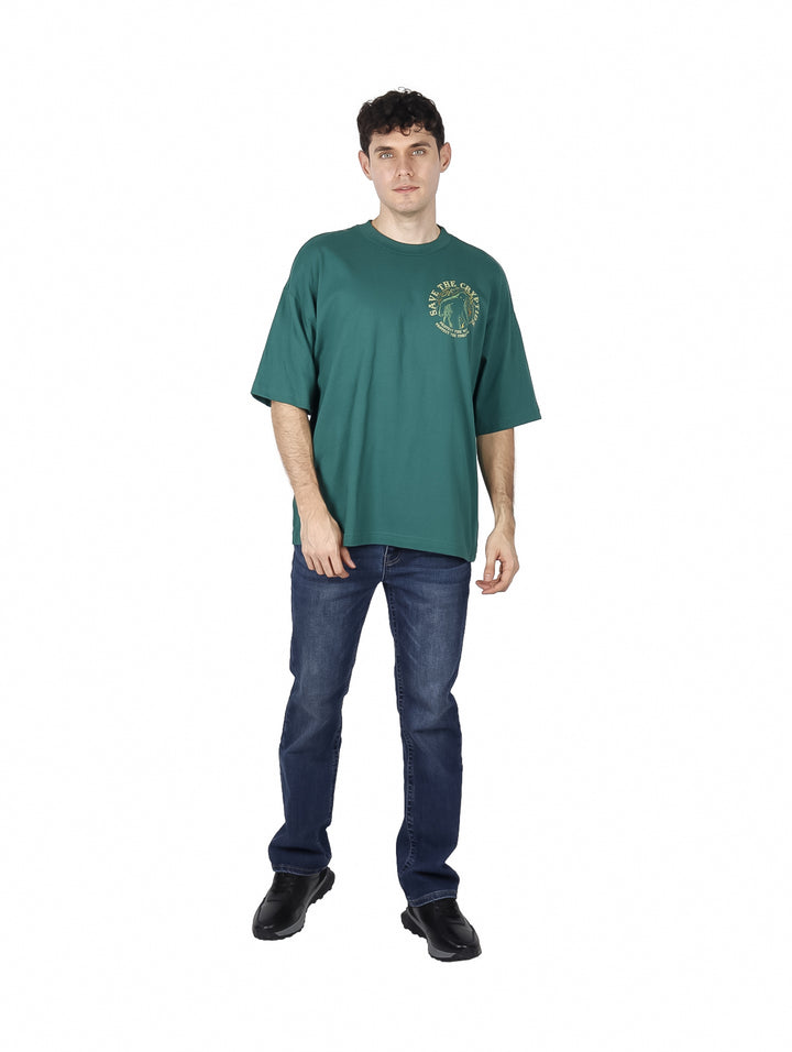 Hombre vistiendo playera oversized color verde botella con estampado amarillo en el pecho.