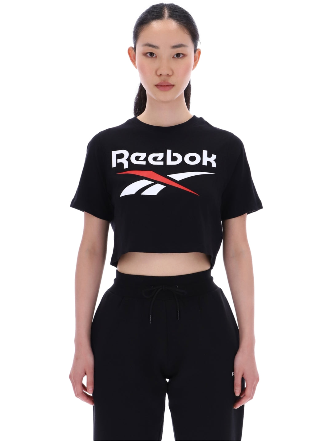Mujer vistiendo camiseta corta negra de Reebok con logo en el pecho y pantalones negros.