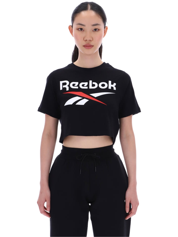 Mujer vistiendo camiseta corta negra de Reebok con logo en el pecho y pantalones negros.