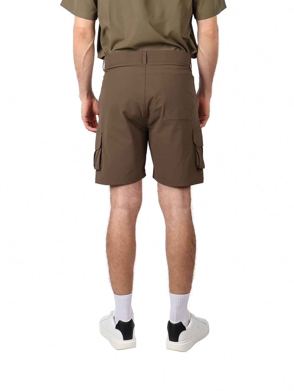Hombre vistiendo bermudas color caf� con bolsillos laterales y calzado blanco sobre fondo blanco.