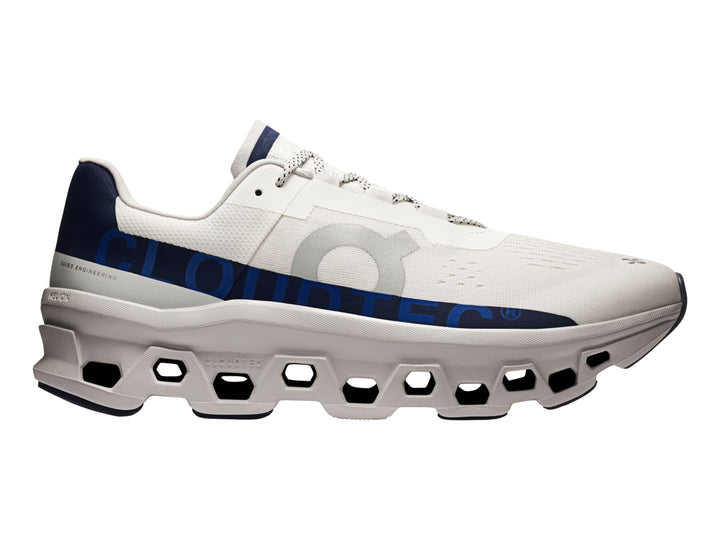 Tenis deportivos ON Cloudmonster en color marfil con detalles en azul marino y suela blanca estructurada.
