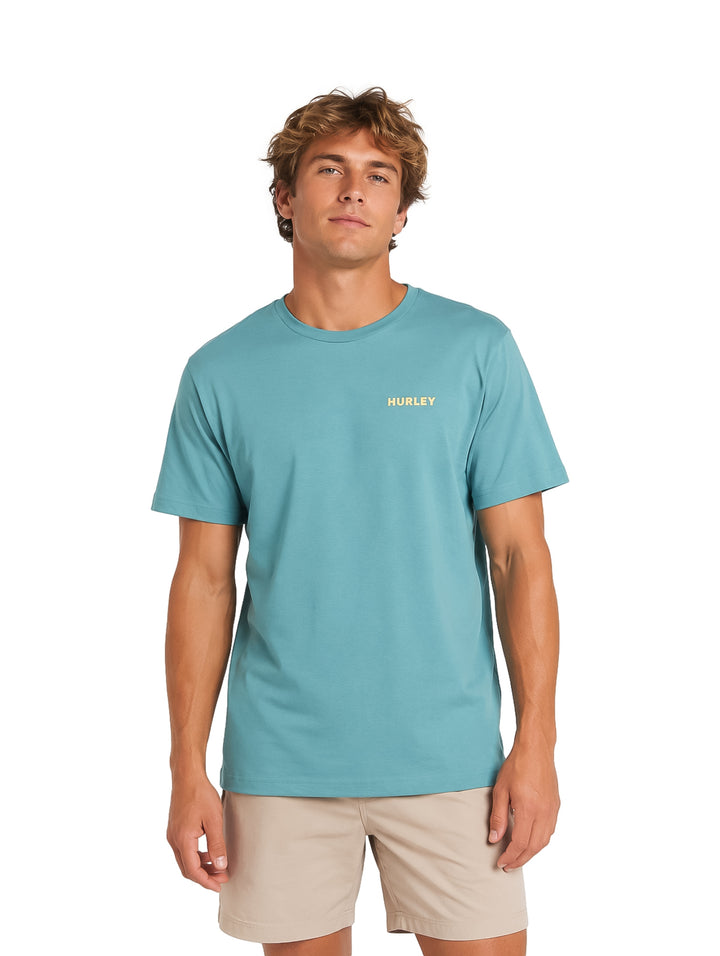Hombre vistiendo playera de manga corta color verde azulado marca Hurley y shorts beige.