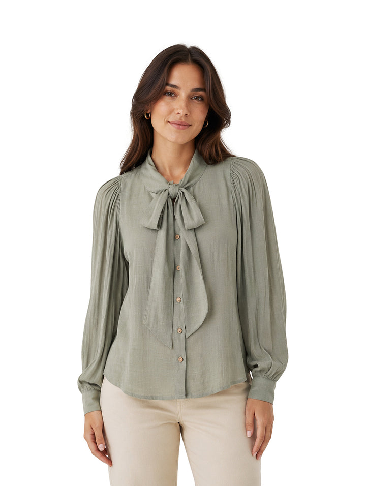 Mujer vistiendo camisa verde de manga larga con lazo en el cuello y pantalones beige.