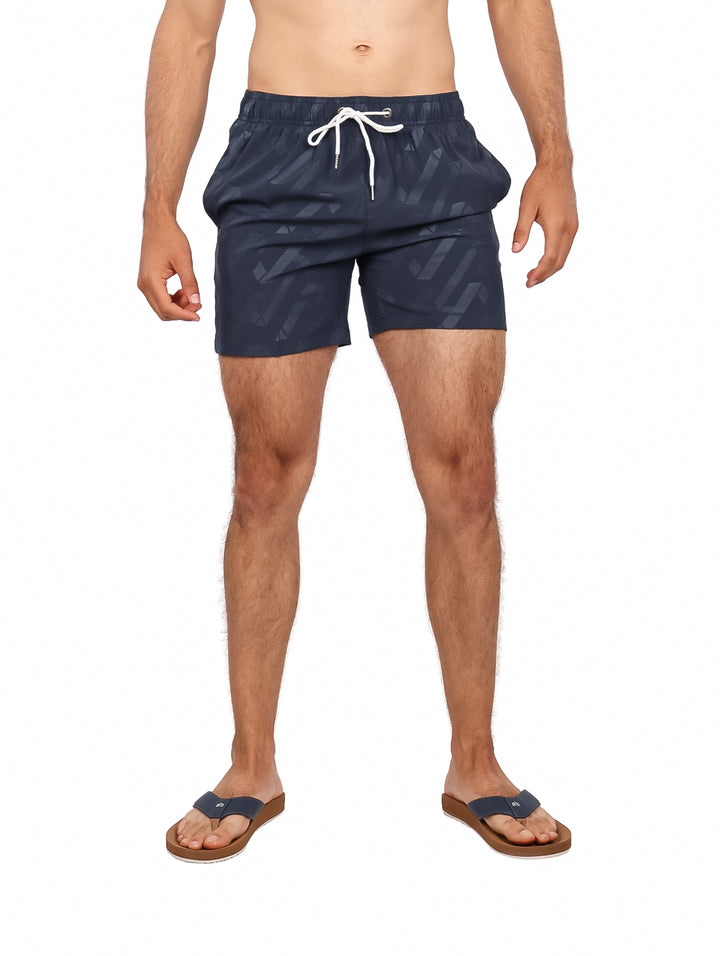 Hombre vistiendo traje de baño corto color azul marino con estampado geométrico y cordón blanco.