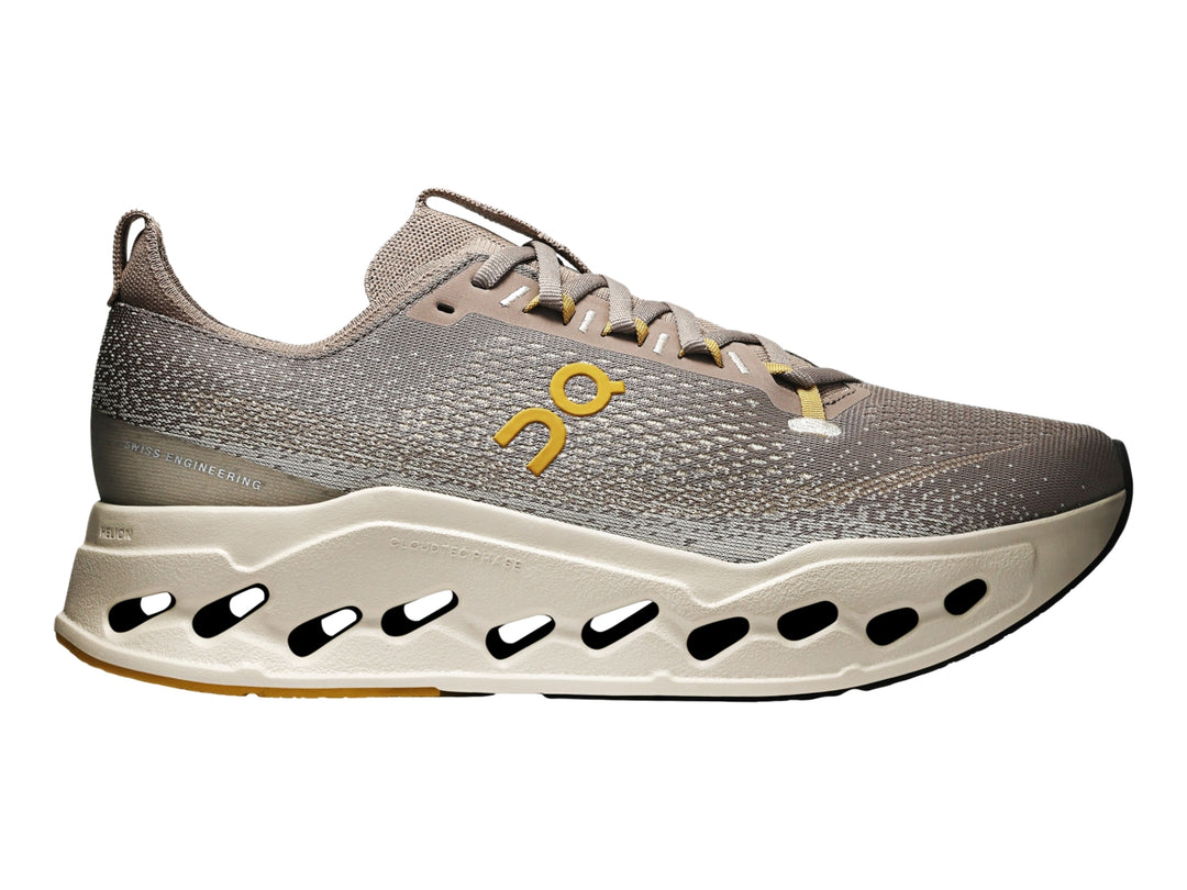 Tenis deportivos On Cloudsurf Max color gris y crema con suela gruesa perforada y logo amarillo.