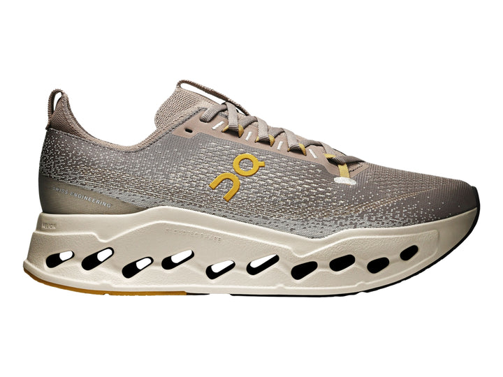 Tenis deportivos On Cloudsurf Max color gris y crema con suela gruesa perforada y logo amarillo.
