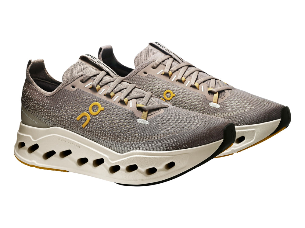 Tenis deportivos On Cloudsurf Max color gris y crema con suela gruesa perforada y logo amarillo.