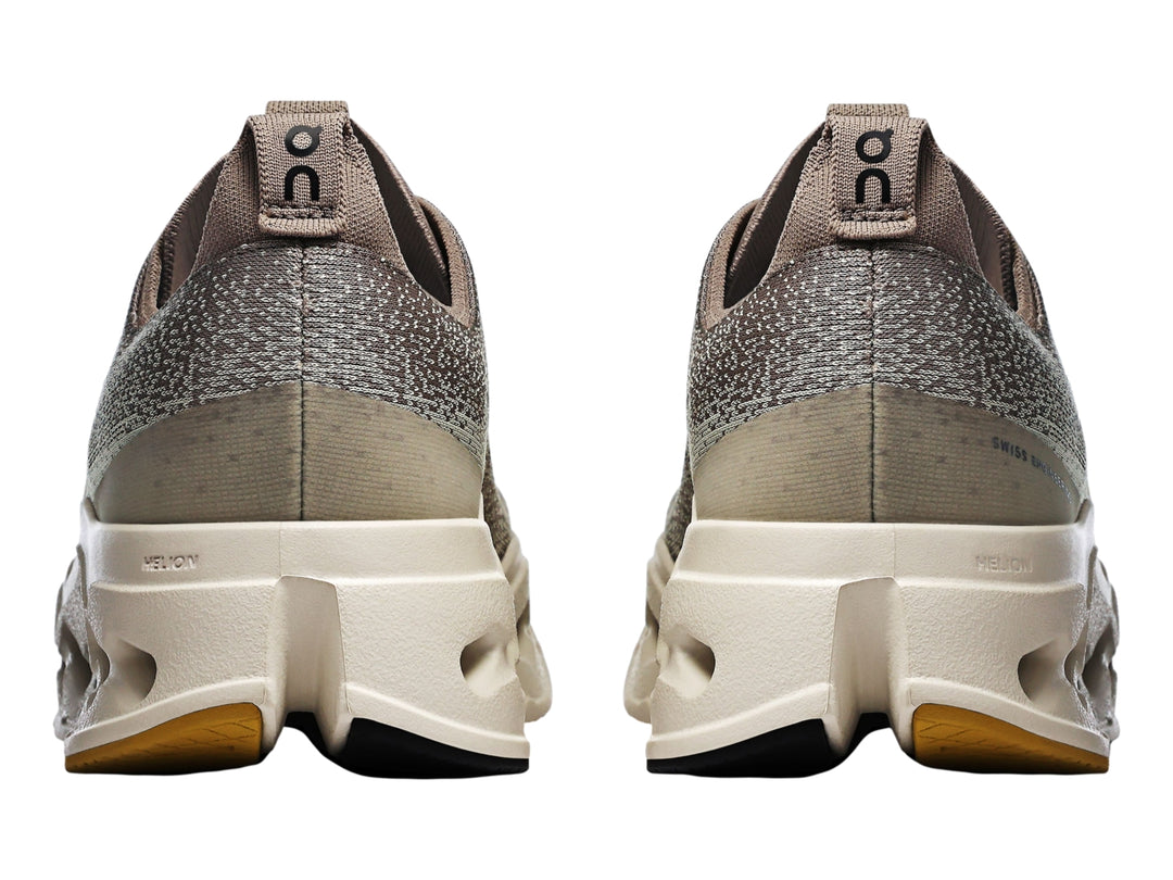 Tenis deportivos On Cloudsurf Max color gris y crema con suela gruesa perforada y logo amarillo.