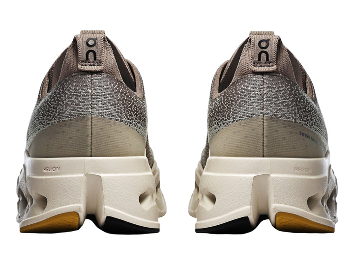 Tenis deportivos On Cloudsurf Max color gris y crema con suela gruesa perforada y logo amarillo.