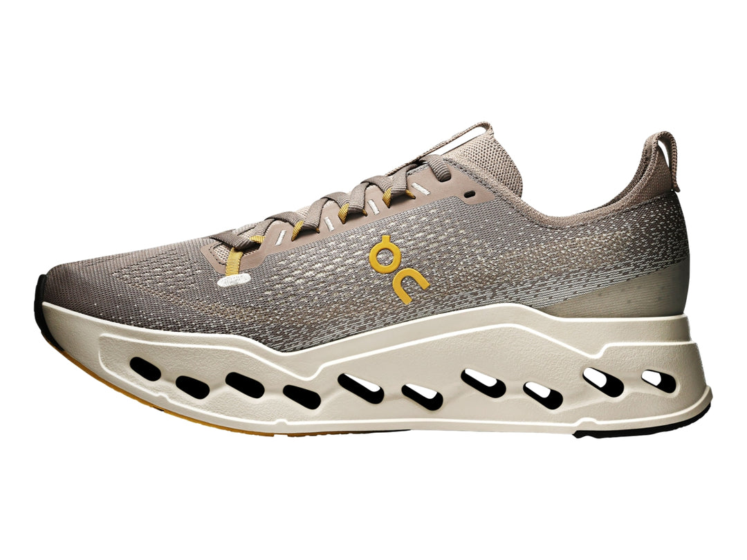 Tenis deportivos On Cloudsurf Max color gris y crema con suela gruesa perforada y logo amarillo.