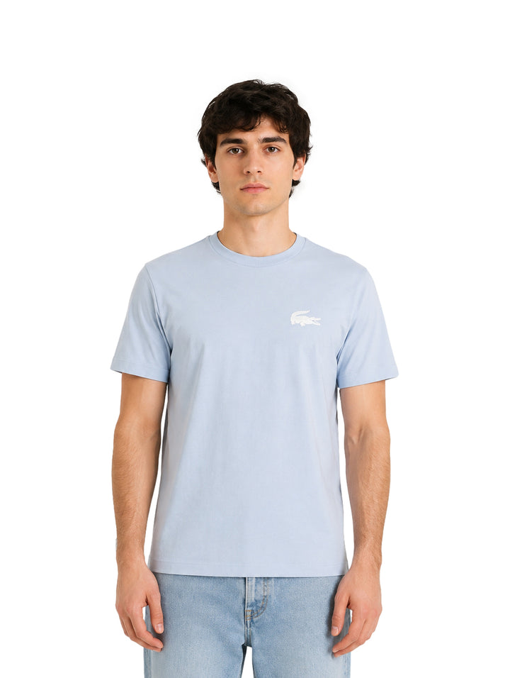 Hombre vistiendo una playera Lacoste de color azul cielo y jeans claros sobre fondo blanco.