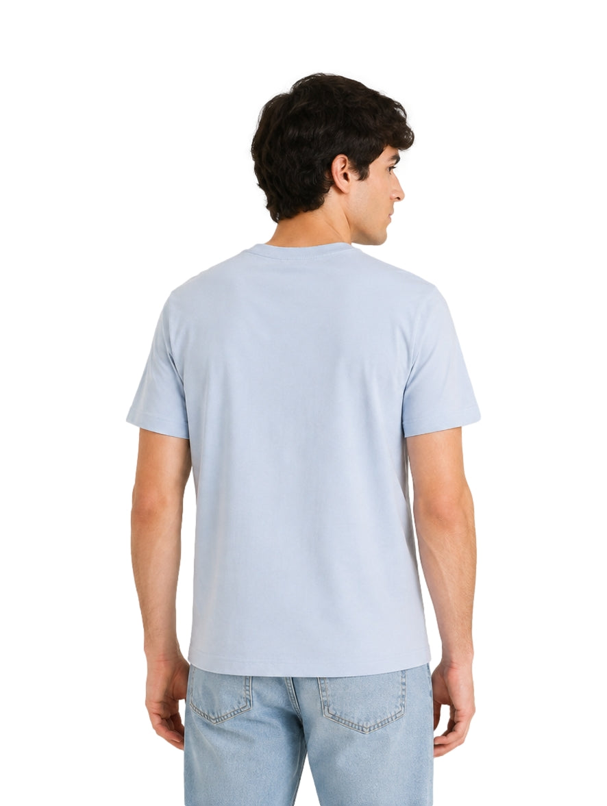 Hombre vistiendo una playera Lacoste de color azul cielo y jeans claros sobre fondo blanco.