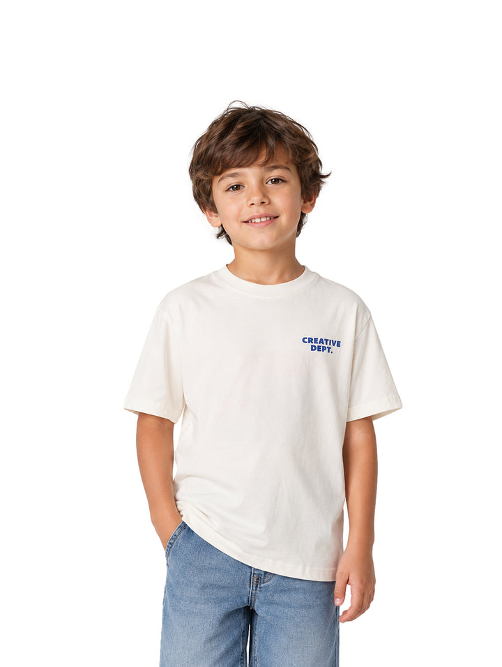 Niño vistiendo una playera blanca de manga corta con texto azul en el pecho y pantalones de mezclilla azules.