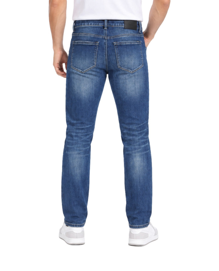 Hombre vistiendo jeans Jeanswest color azul medio stone con calzado blanco sobre fondo blanco.