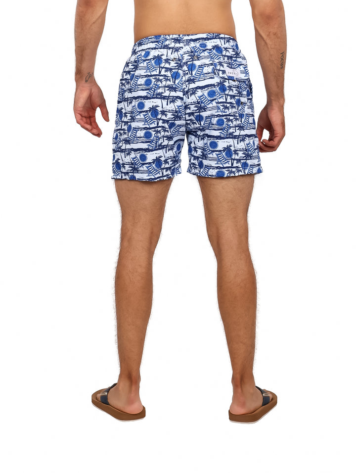 Hombre vistiendo traje de baño corto estampado en color azul marino y blanco con diseño de palmeras.