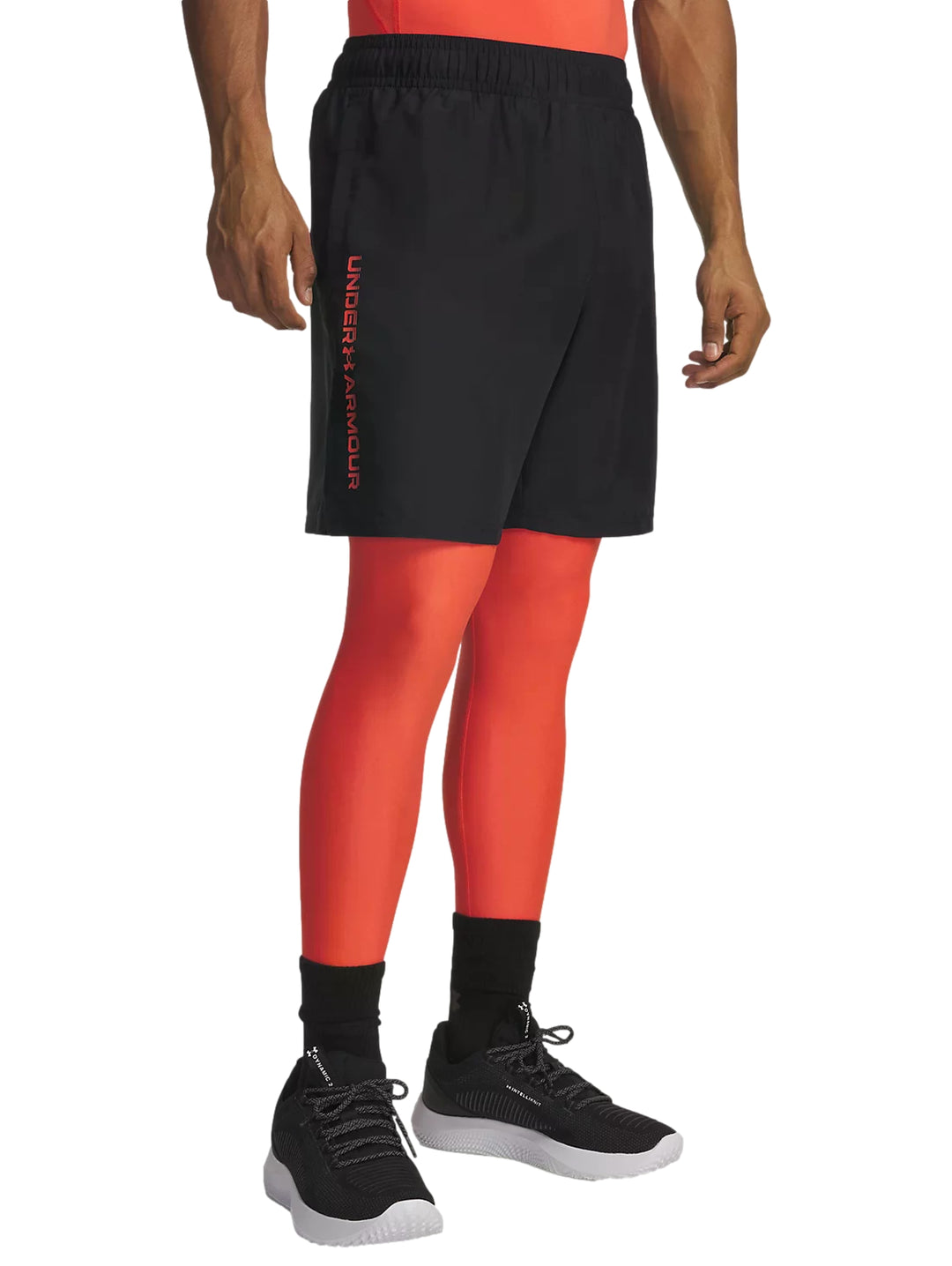 Hombre vistiendo shorts deportivos negros de Under Armour sobre mallas naranjas y tenis negros.