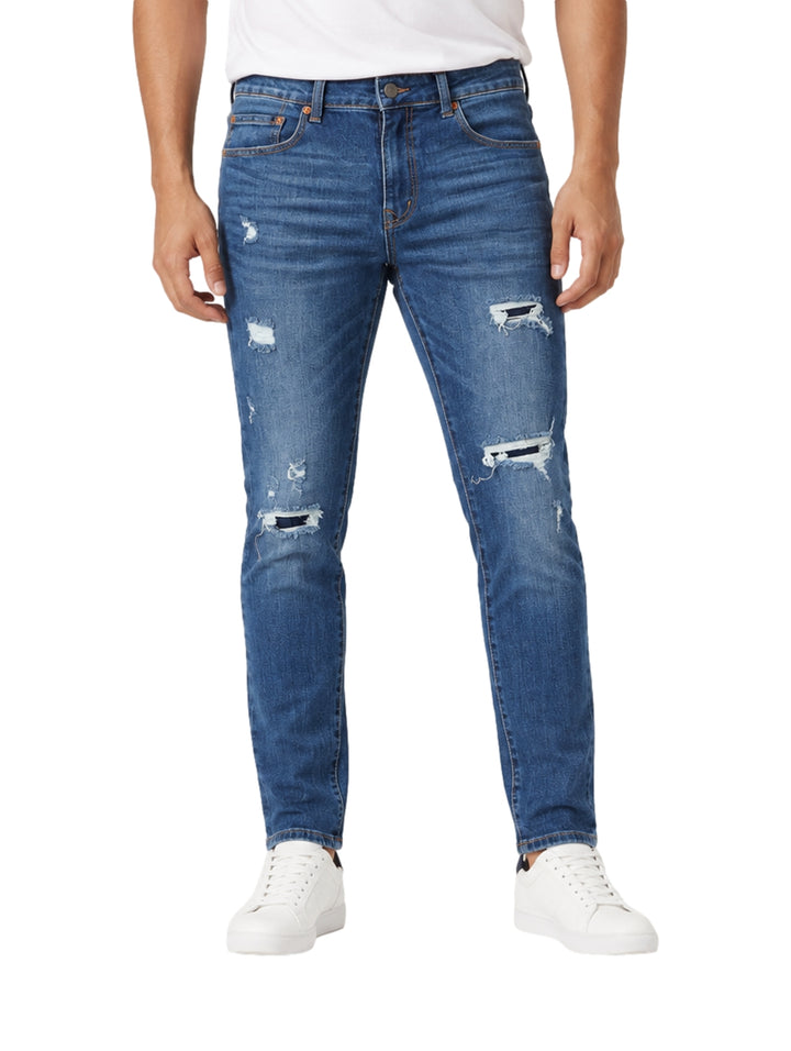 Hombre usando jeans de mezclilla azul medio con efecto roto y tenis blancos.