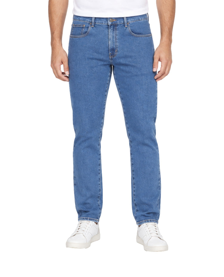 Hombre vistiendo jeans OGGI color stone blue con tenis blancos sobre fondo blanco.