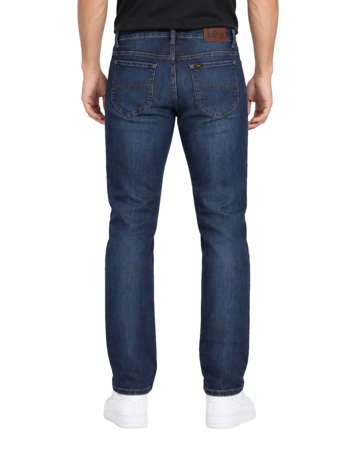 Hombre vistiendo jeans slim fit Lee color azul stone con tenis blancos sobre fondo blanco.