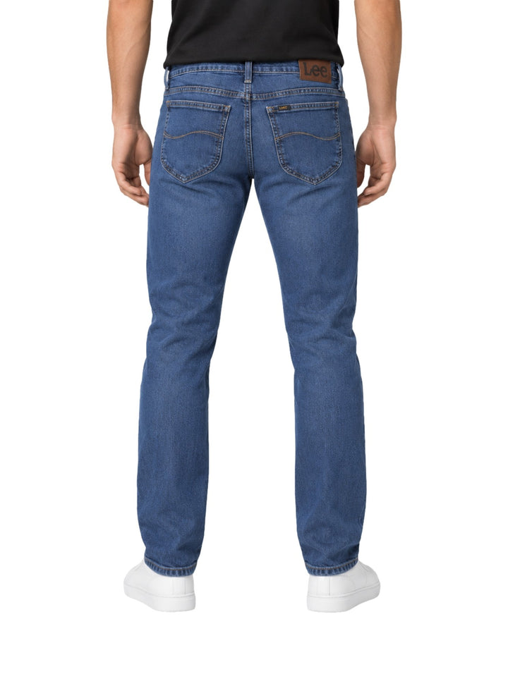 Hombre vistiendo jeans Lee de color azul con calzado blanco sobre fondo blanco.
