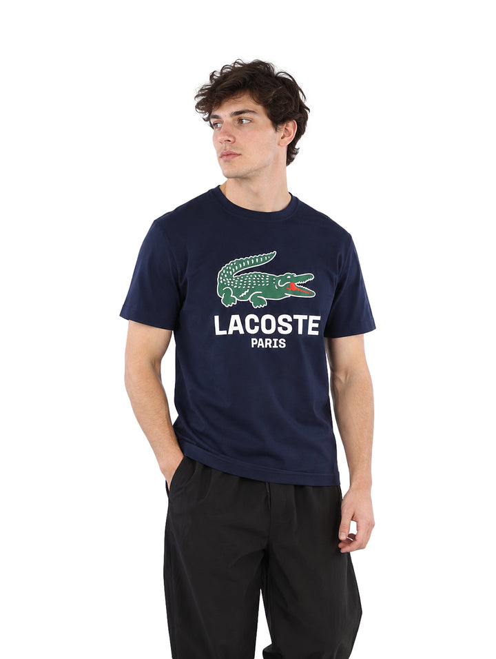 Hombre vistiendo playera Lacoste color navy con logotipo frontal y pantalones negros.