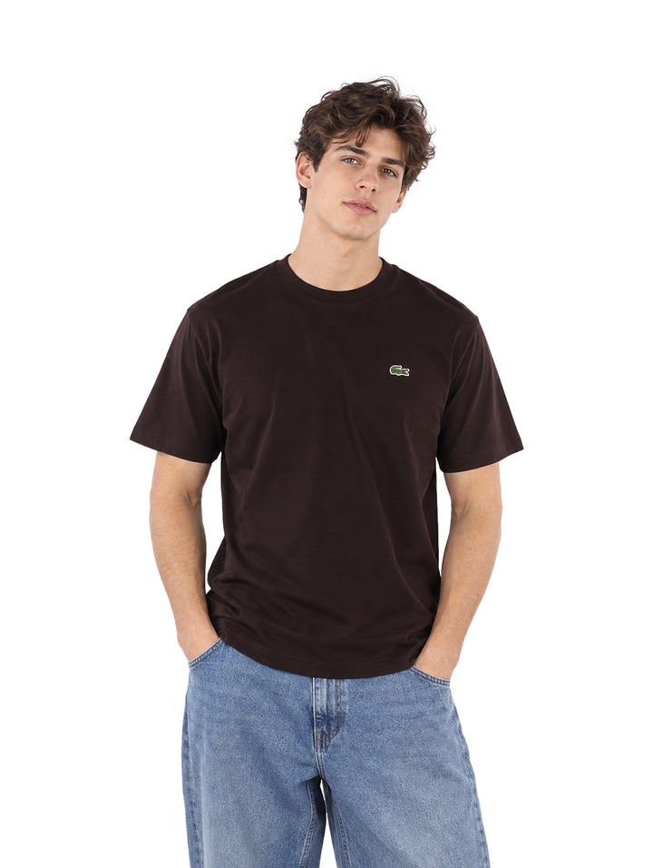 Hombre vistiendo una playera Lacoste de color café y pantalones de mezclilla azules sobre fondo blanco.
