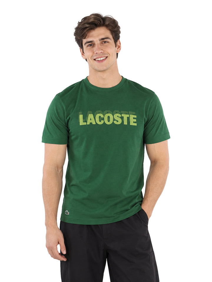 Hombre vistiendo playera Lacoste color verde de manga corta con texto frontal y shorts negros.