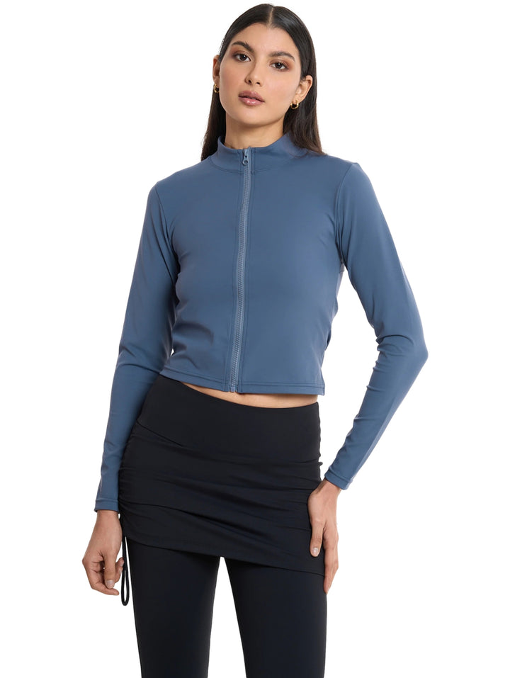 Mujer vistiendo chaqueta deportiva corta color acero con cierre frontal y leggings negros.