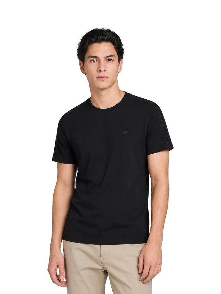 Hombre vistiendo una playera negra de manga corta DKNY y pantalones beige sobre fondo blanco.