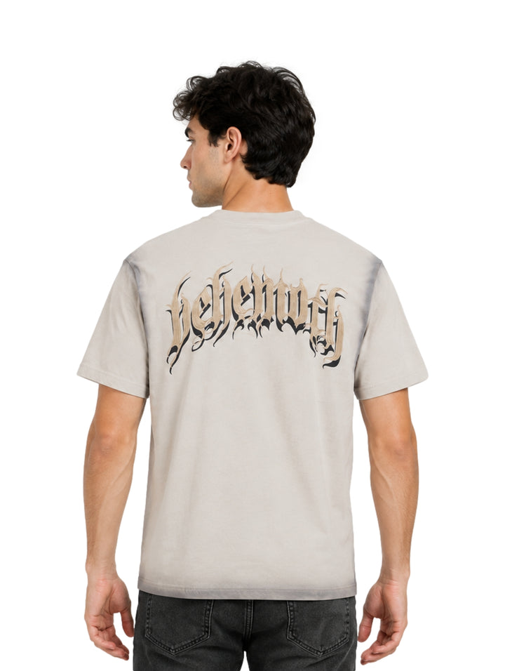 Hombre vistiendo playera beige de manga corta con detalles degradados en gris y estampado gótico central.