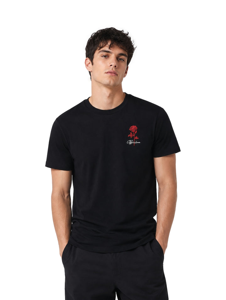 Hombre vistiendo playera negra de manga corta con un pequeño estampado de rosa roja en el pecho.