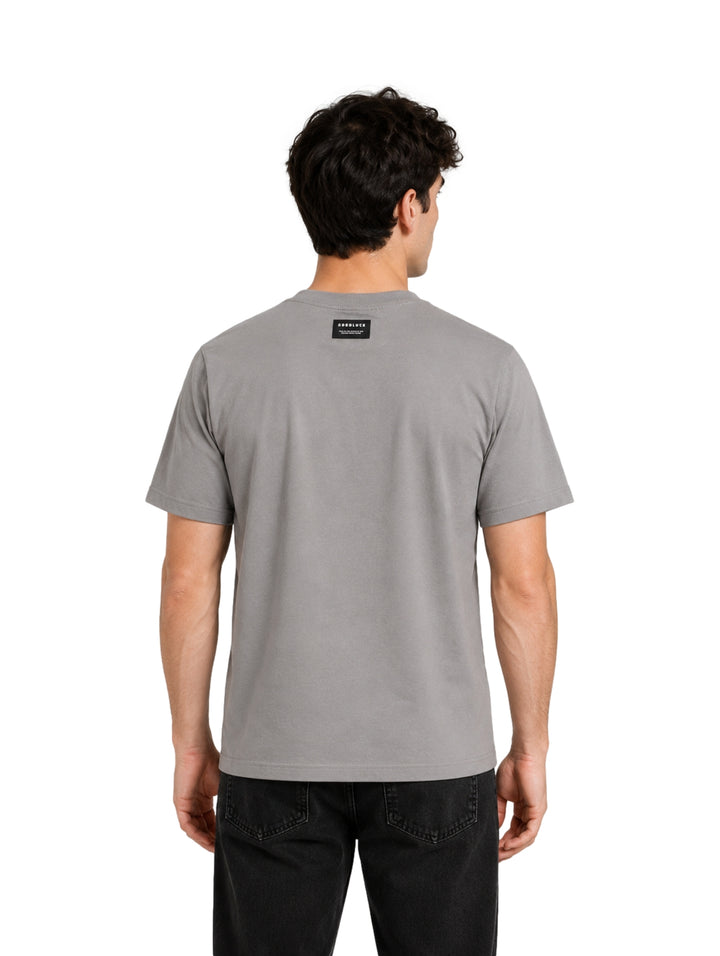 Hombre vistiendo una playera gris de manga corta con texto negro en el pecho.