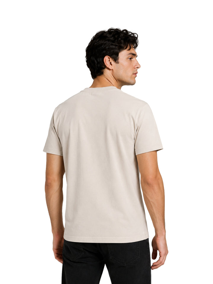 Hombre vistiendo playera de color hueso de manga corta con logotipo pequeño en el pecho.
