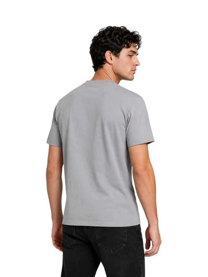 Hombre vistiendo una playera de color gris de manga corta con un pequeño logotipo negro en el pecho.