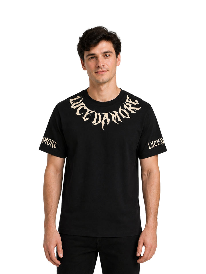 Hombre vistiendo playera negra de manga corta con letras góticas en color beige en el pecho y mangas.