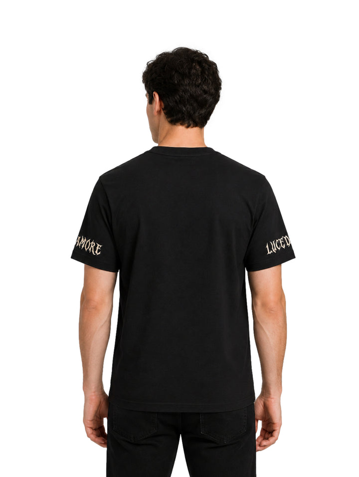 Hombre vistiendo playera negra de manga corta con letras góticas en color beige en el pecho y mangas.