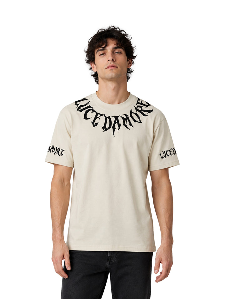 Hombre vistiendo playera color beige de manga corta con diseño de letras góticas negras en el cuello y mangas.